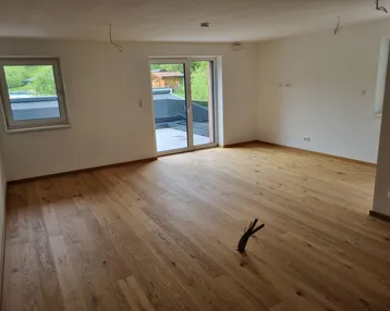 Helles, leeres Zimmer mit Holzboden und Zugang zu einer Terrasse, umgeben von grüner Natur.