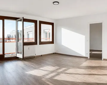Helles, leeres Wohnzimmer mit großen Fenstern, Laminatboden und Zugang zu einem Balkon.