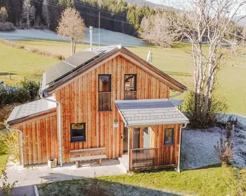 Modernes Holzhaus mit Photovoltaikanlage auf dem Dach, umgeben von grünen Wiesen und Bergen.