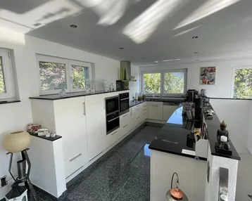Moderne Küche mit weißen Schränken, dunkler Arbeitsplatte und großformatigen Fliesen. Helle Fenster bieten Blick ins Grüne.