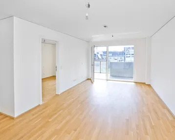 Helles, leeres Wohnzimmer mit Parkettboden und Zugang zu einem Balkon.