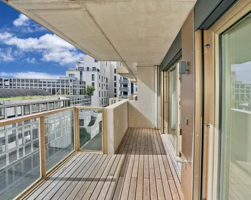 Geräumiger Balkon mit Holzboden und Glasgeländer, bietet einen Blick auf die umliegenden Gebäude.