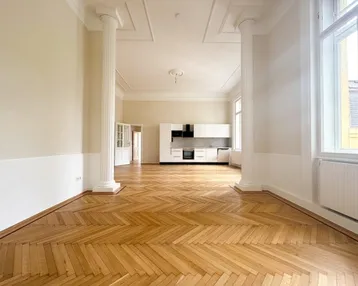 Geräumiger, lichtdurchfluteter Raum mit Fischgrätparkett, hohen Decken, Säulen und integrierter moderner Küche.