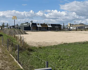 Blick auf ein McDonald's Restaurant mit einem großen Schotterparkplatz und angrenzenden Grünflächen unter blauem Himmel.