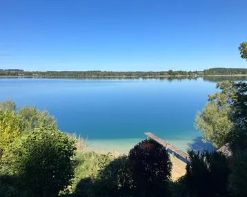Blauer See mit kleinem Holzsteg und üppiger Vegetation im Vordergrund unter klarem Himmel.