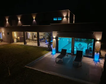 Moderne Villa bei Nacht mit beleuchteter Terrasse, Garten und Poolbereich.
