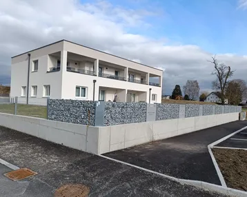 Modernes Mehrfamilienhaus mit heller Fassade, Balkonen und Gabionenmauer im Außenbereich.