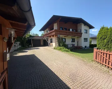 Traditionelles Haus mit Balkonen, gepflasterter Auffahrt und Carport unter blauem Himmel.