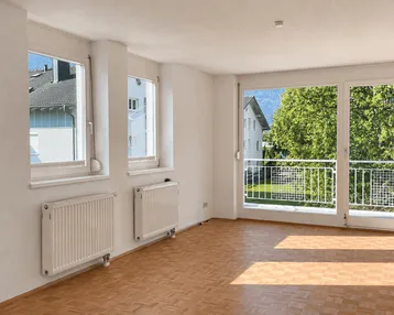 Helles Wohnzimmer mit Parkettboden, großen Fenstern und Zugang zu einem Balkon mit Grünblick.