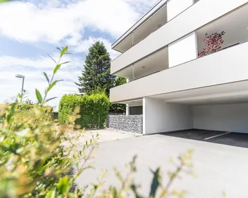 Moderne Wohnanlage mit Balkonen und überdachtem Parkplatz, umgeben von grüner Bepflanzung.