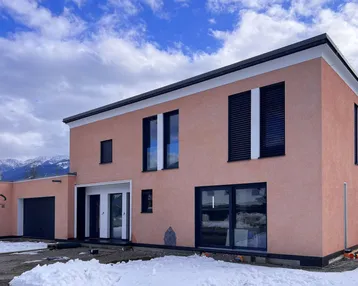 Modernes Einfamilienhaus mit rosafarbener Fassade und dunklen Fensterläden vor verschneiter Bergkulisse.