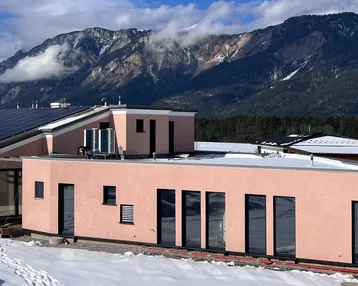 Modernes Haus mit rosafarbener Fassade, Flachdach und Photovoltaikanlage vor verschneiter Berglandschaft.