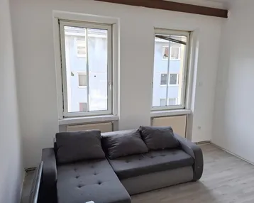 Helles Wohnzimmer mit grauem Ecksofa, Laminatboden und Blick auf gegenüberliegende Gebäude.