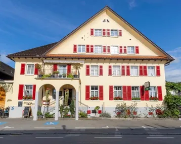 Traditionelles Gasthaus mit gelber Fassade, roten Fensterläden und Balkonen unter blauem Himmel.