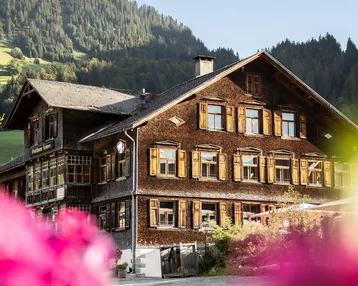 Traditionelles Holzhaus in den Bergen mit grüner Umgebung und blühenden Pflanzen im Vordergrund.