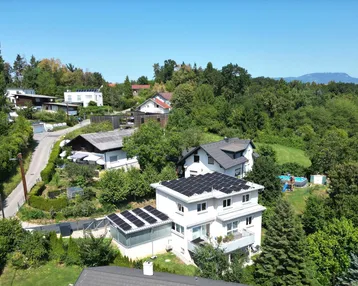 Luftaufnahme eines Wohnhauses mit Solaranlage und umliegender grüner Landschaft unter blauem Himmel.