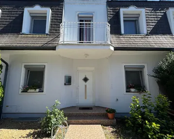 Weiße Hausfassade mit Mansarddach, kleinen Balkonen und gepflegtem Vorgarten unter blauem Himmel.