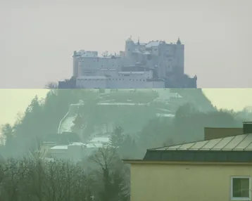 Fernblick auf eine historische Burg auf einem Hügel, umgeben von Bäumen und Nebel.