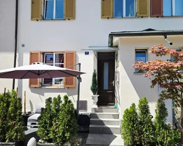 Vorderansicht eines Reihenhauses mit Eingangsbereich, Fensterläden und kleinen Zierbäumen.