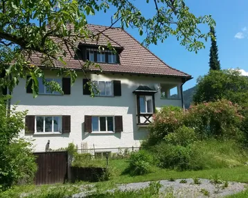 Traditionelles Haus mit Satteldach, umgeben von üppigem Grün und Bäumen unter blauem Himmel.