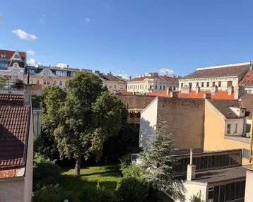 Blick auf einen grünen Innenhof mit Bäumen und umliegenden Gebäuden unter blauem Himmel.