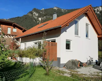 Seitenansicht eines Einfamilienhauses mit weißer Fassade, rotem Dach und Bergblick.
