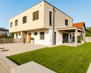 Modernes Einfamilienhaus mit heller Fassade, gepflegtem Rasen und gepflastertem Vorplatz.