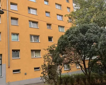 Mehrfamilienhaus mit orangefarbener Fassade, vielen Fenstern und einem Gründach, umgeben von Bäumen und Rasen.