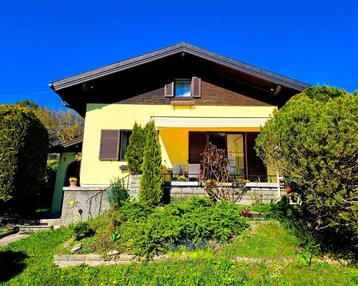 Gelbes Einfamilienhaus mit dunklem Dach, umgeben von einem gepflegten Garten und einer möblierten Terrasse.