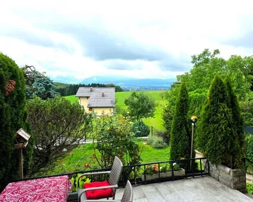 Terrasse mit Gartenmöbeln und weitem Blick über die grüne Landschaft und Berge unter bewölktem Himmel.