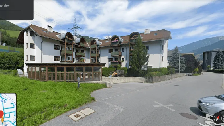 Mehrfamilienhaus mit Balkonen, umgeben von Grünflächen und Berglandschaft unter blauem Himmel.