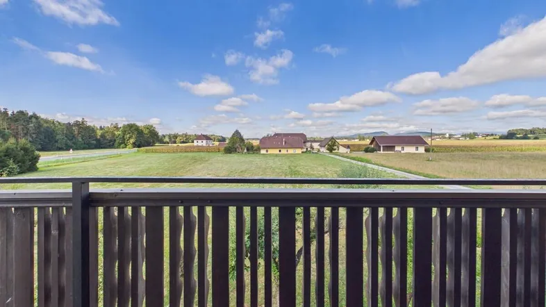 Blick vom Balkon auf die weite, grüne Landschaft und umliegende Felder unter blauem Himmel.