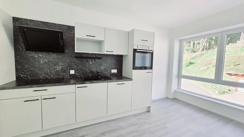 Moderne Einbauküche mit weißen Schränken, dunkler Arbeitsplatte und großem Fenster mit Blick ins Grüne.