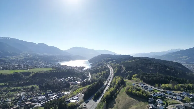 Weitläufige Luftaufnahme mit strahlendem Sonnenschein, einem See, Bergen und einer Autobahn in der Ferne.