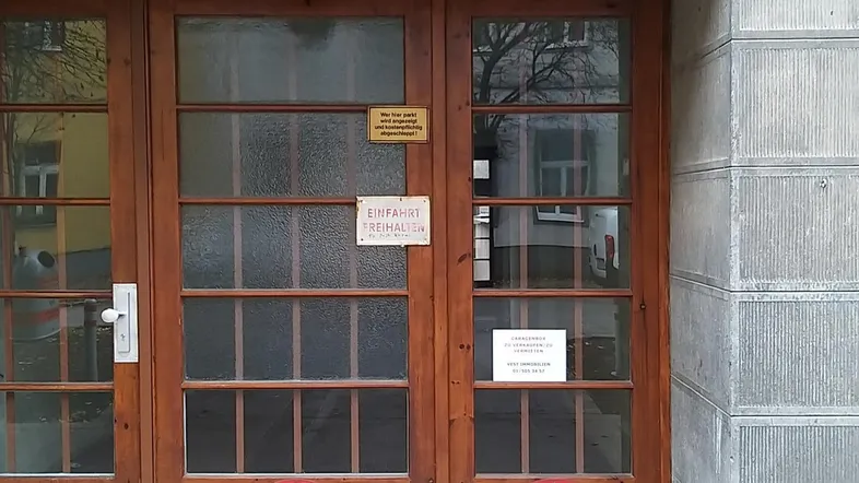 Eingangsbereich eines Gebäudes mit Holztür, Hausnummer 16 und Hinweisschildern auf dem Boden.