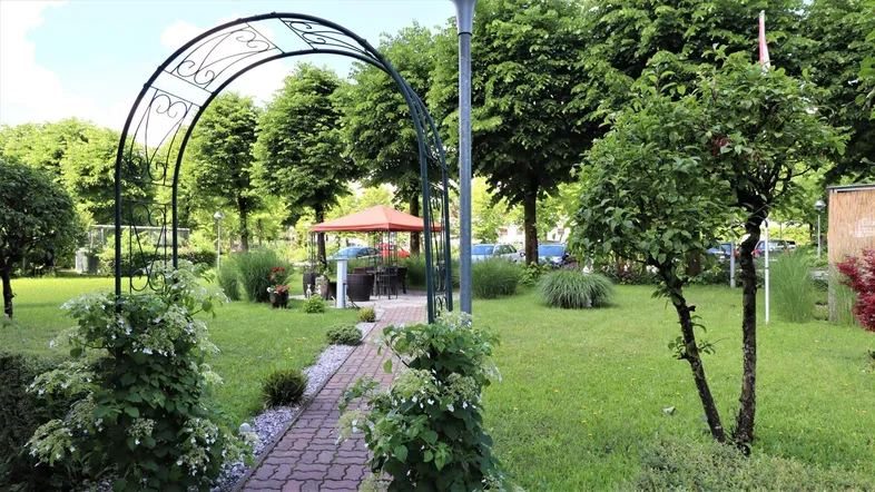 Grüner Gartenbereich mit einem geschwungenen Metallbogen und einem Pavillon im Hintergrund.