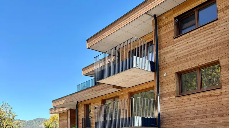 Modernes Gebäude mit Holzfassade, mehreren Balkonen und Bergblick unter blauem Himmel.