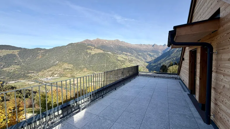 Große geflieste Terrasse mit weitem Bergblick und Holzfassade des Neubaus unter blauem Himmel.