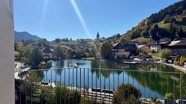Sonniger Balkon mit atemberaubendem Blick auf einen ruhigen See, ein charmantes Dorf und die majestätischen Berge.
