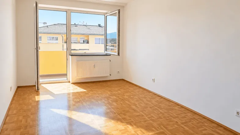 Helles, leeres Zimmer mit Parkettboden und Zugang zu einem kleinen Balkon mit Blick auf die Umgebung.