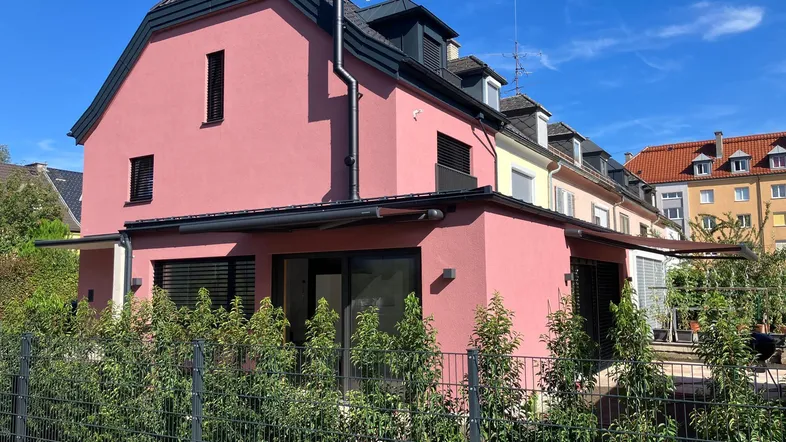 Modernes Reihenhaus mit rosafarbener Fassade, dunklem Dach und grüner Hecke im Vorgarten.