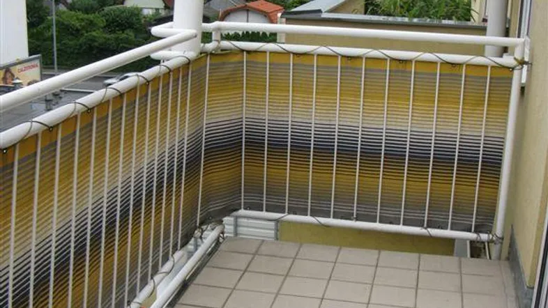 Balkon mit Fliesenboden, weißem Geländer und gelb-schwarzem Sichtschutz, bietet einen Blick auf die Umgebung.