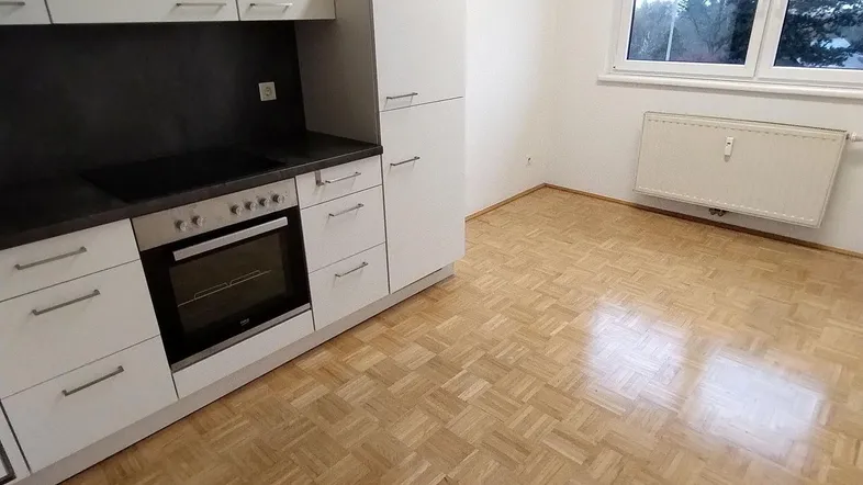 Moderne Küchenzeile mit weißen Schränken, dunkler Arbeitsplatte und Parkettboden in einem hellen Raum.