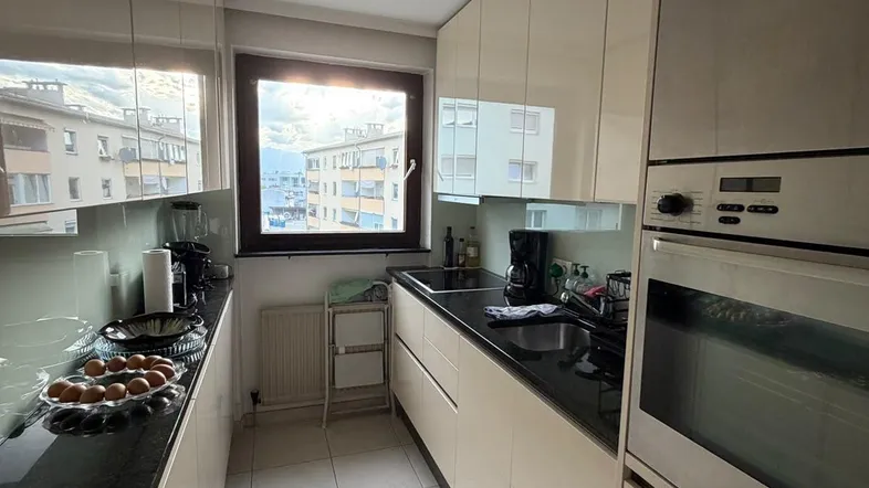 Helle, schmale Küche mit beigen Hochglanzfronten, dunkler Arbeitsplatte und Fensterblick.