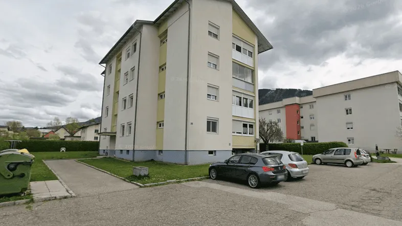 Mehrfamilienhaus mit hellgelber Fassade, Parkplätzen und Grünflächen unter bewölktem Himmel.