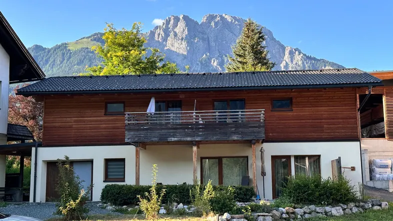 Modernes Haus mit Holzfassade und Balkon, umgeben von Grünflächen mit beeindruckendem Bergblick.