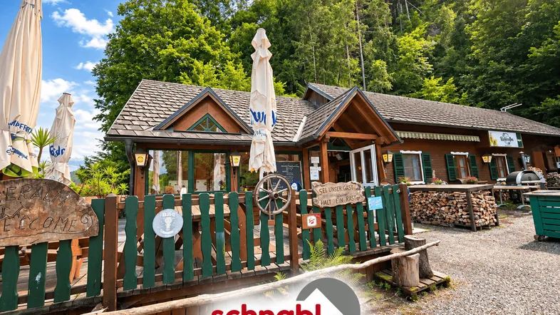Rustikales Holzgebäude mit Außenterrasse und Sonnenschirmen, umgeben von grünen Bäumen und Bergen.