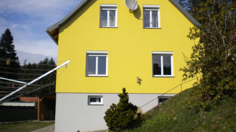 Gelbes Einfamilienhaus mit Satteldach und Rollläden vor bewaldetem Hintergrund.