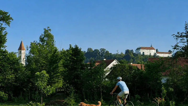 Weitläufige grüne Landschaft mit Radfahrer und Hund, im Hintergrund ein historisches Gebäude und eine Kirche unter blauem Himmel.