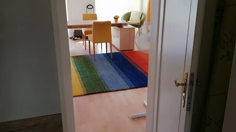 Blick durch eine offene Tür in einen hellen Raum mit Holzboden, bunten Teppichen und gelben Stühlen.